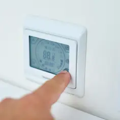 Dłoń naciska przycisk białego termostatu z cyfrowym wyświetlaczem.